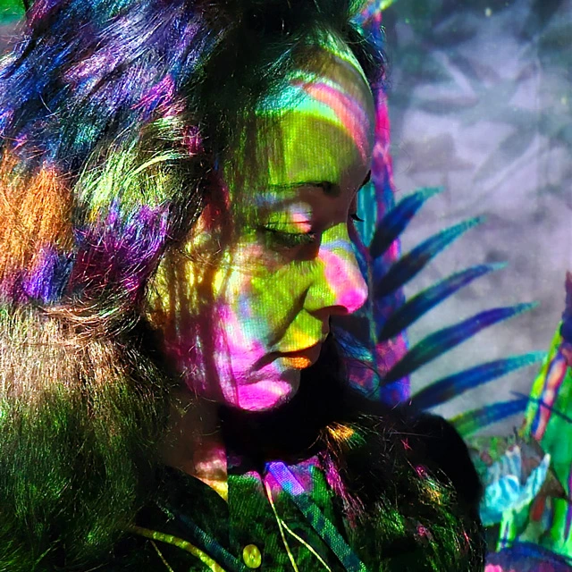 Anna Nogaré’s face close up looking down with multicolored light and foliage projections on her face and background.