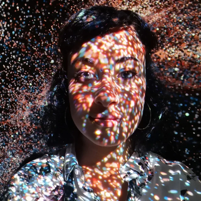 Anna Nogaré facing the camera with colorful dot projections on her face and shirt, against a dark background.