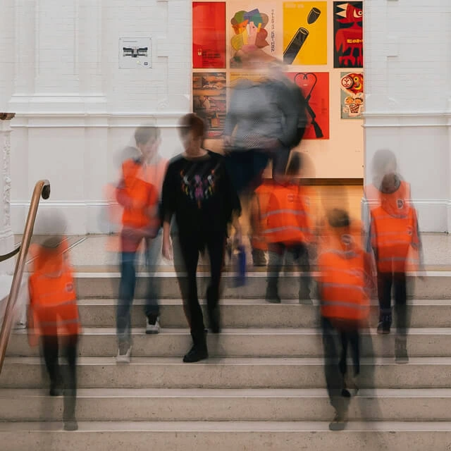 A long-exposure shot of a crowd moving down white stone steps in a museum, with vibrant graphic design posters on the wall behind them.