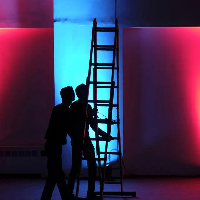 Dramatic silhouette of a production crew and a wooden ladder against glowing blue and magenta walls.