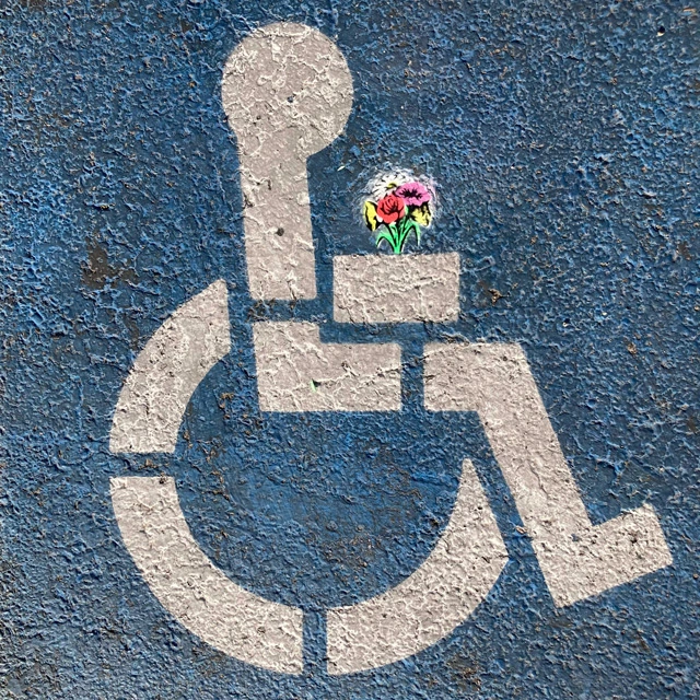 Stylized wheelchair symbol painted on asphalt, with a small bouquet of flowers placed in the figure’s hands.