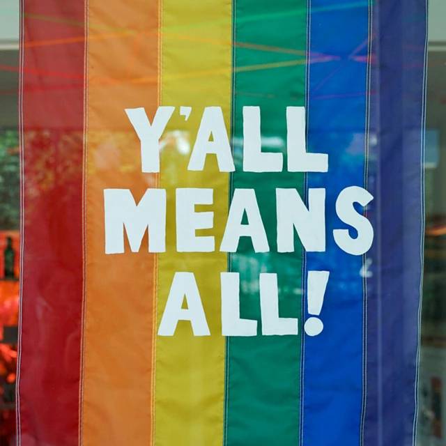 Rainbow-striped Pride flag with bold white text reading “Y’all Means All!” hanging inside a window.