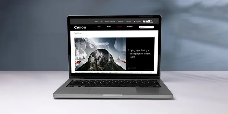 A front view of an open laptop sitting on a white surface against a soft grey-blue backdrop. The screen displays the Canon Professional Network website, showcasing a technical article titled “Flying high: filming an air display with the EOS C700” with a photo of a pilot inside a cockpit.
