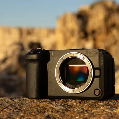 A Canon mirrorless camera body resting on a rough, textured stone surface outdoors during the warm light of golden hour. The camera lens is removed, clearly exposing the reflective internal image sensor, with a blurred rocky landscape in the background.