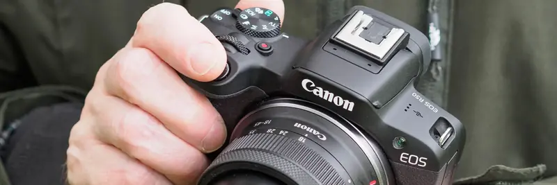 A close-up shot of a hand securely holding a black Canon EOS R50 mirrorless camera fitted with an RF-S 18-45mm lens. The user’s index finger is poised over the shutter button, and the thumb rests near the top control dials, set against the green fabric of a jacket.