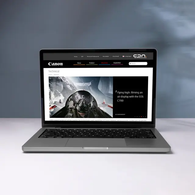 A front view of an open laptop sitting on a white surface against a soft grey-blue backdrop. The screen displays the Canon Professional Network website, showcasing a technical article titled “Flying high: filming an air display with the EOS C700” with a photo of a pilot inside a cockpit.
