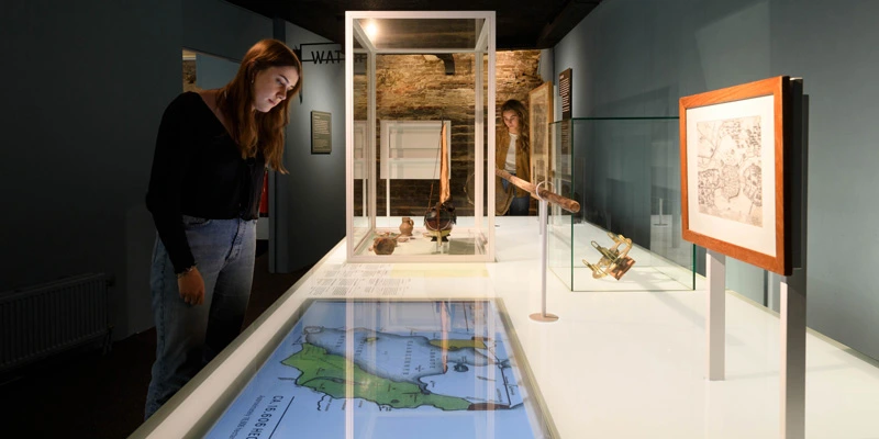 Museum visitor exploring a display case containing navigation instruments, maps, and archaeological artifacts, with a touch screen integrated into the surface.
