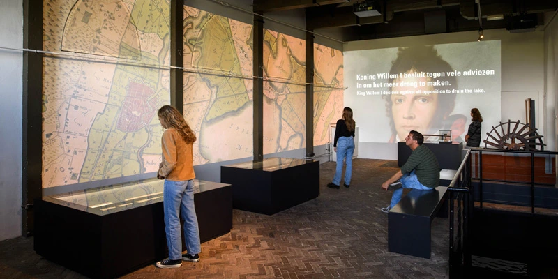 Panoramic view of the gallery featuring an immersive wall projection of historical maps and a portrait of King Willem I, with visitors engaging with the exhibits.