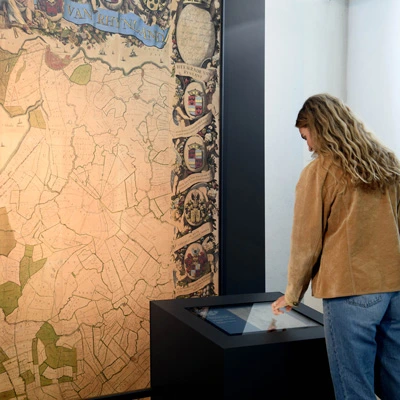 Woman interacting with a digital touchscreen in front of a large decorative map, using the interface to explore historical information.