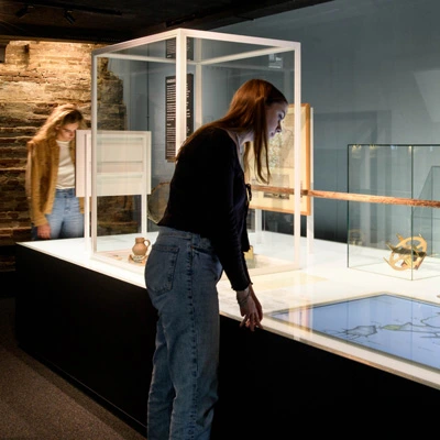 Two visitors interacting with a long display table featuring historical navigation tools, ceramic artifacts, and a digital map interface, in a gallery combining exposed brick and museum lighting.