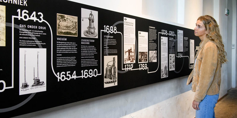 Woman examining a large wall-mounted visual timeline that traces the development of steam technology from 1643 onward, featuring historical texts and illustrations.