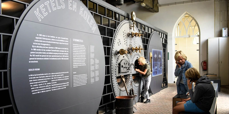 Child peering into a reconstructed boiler installation, while two other children in the foreground use audio handsets to explore the exhibit in the “Boiler Room” section.