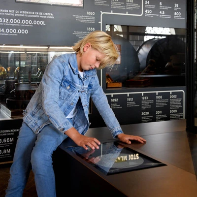 Child interacting with a horizontal touchscreen table, placed in front of a large infographic panel showing engineering data and historical timelines of the pumping station.