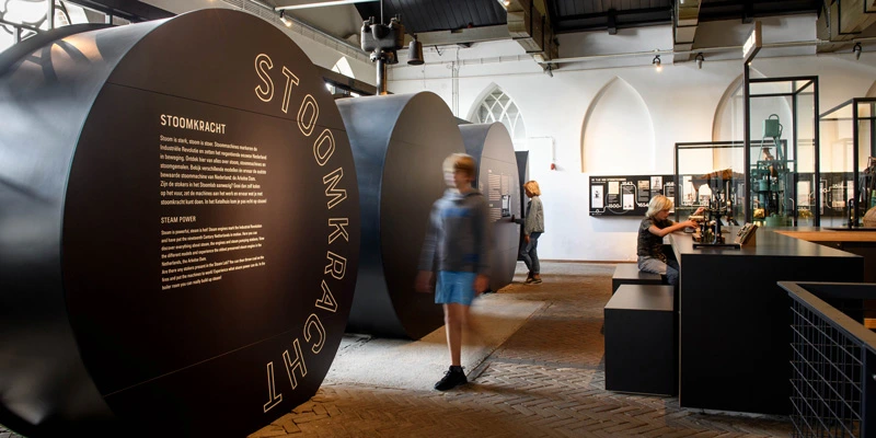 Children interacting with tall, black cylindrical structures that present historical information about steam power, surrounded by other exhibits and timeline graphics.
