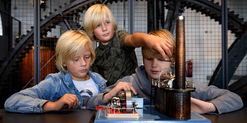 Three children closely examining a hands-on model of a steam engine, focusing on its moving mechanical parts.