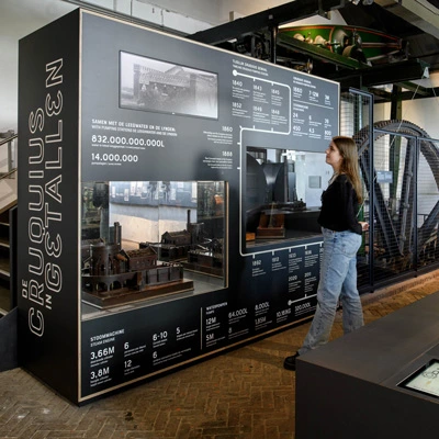 A young woman observing a detailed infographic display with historical data, diagrams, and scale models related to the Cruquius pumping station and its steam engines.