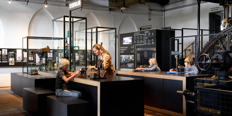 Children seated at a long interactive counter, experimenting with steam engine models, assisted by a woman under a “Stoomlab” sign in the background.