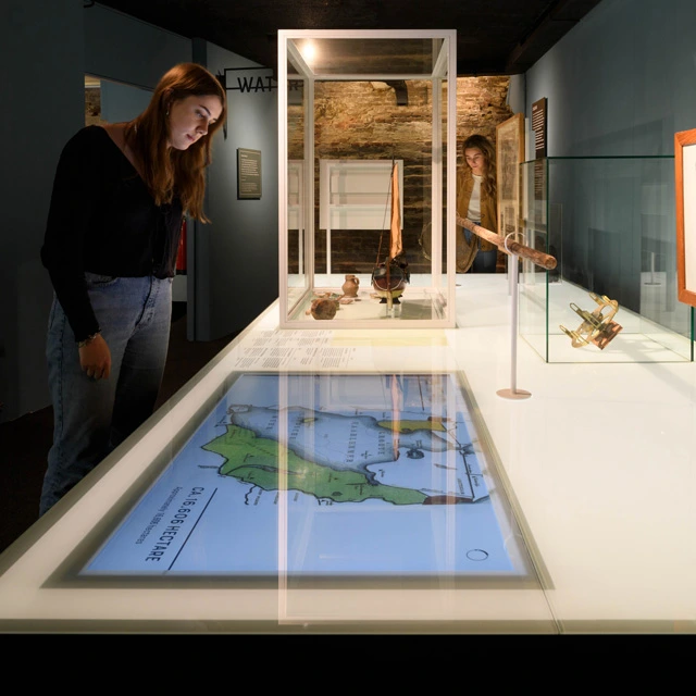 Museum visitor exploring a display case containing navigation instruments, maps, and archaeological artifacts, with a touch screen integrated into the surface.