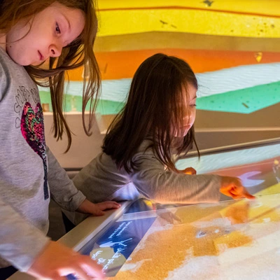 Two young girls interacting with a sand-textured touchscreen interface, part of a digital digging game that simulates uncovering buried artifacts in different soil layers.