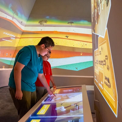 Two teenagers engaging with a large horizontal touchscreen as part of an interactive archaeological game, set within a gallery where colorful stratigraphic layers are projected on the walls to simulate excavation contexts.