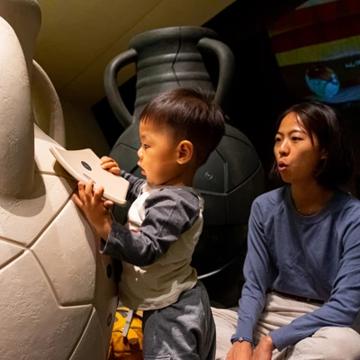 A toddler inserting a puzzle piece into a large interactive ceramic-alike amphora, part of a hands-on archaeological activity, while an adult woman looks on.