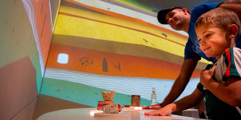 Boy and father engaging with an interactive table game where participants place their hands over specific object replicas to trigger animated projections on the wall, revealing excavation layers and archaeological timelines.