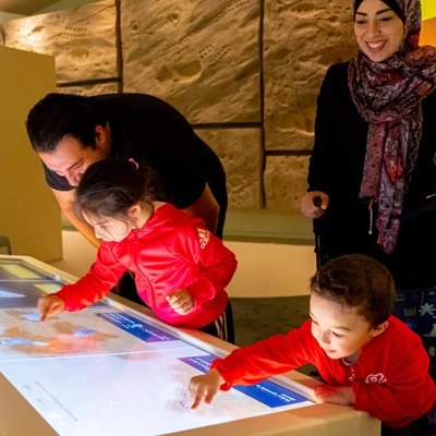 Children using a multi-user touchscreen to uncover virtual artifacts in a digging-themed game, while parents look on, in a gallery designed to simulate a layered archaeological dig site.