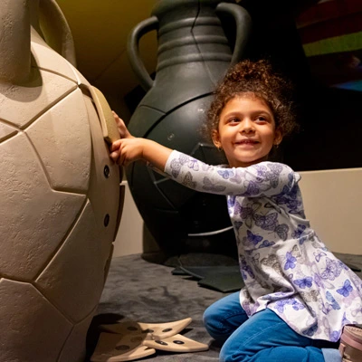 Young girl smiling while playing with a tactile puzzle on a large amphora replica, part of an interactive installation that invites children to reconstruct broken archaeological artifacts.