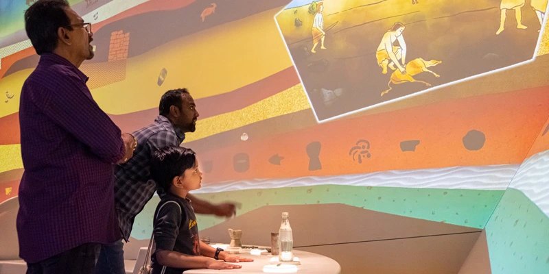 A child interacting with replicas of ancient artifacts on a round table; wall projections above respond with animated historical scenes and layers representing excavation contexts. Two adult men are observing the game.