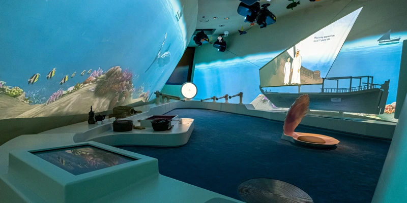 Wide view of a gallery with immersive underwater projections covering the walls; traditional fishing tools, props and interactive screens are arranged on display, and a large stylized pearl shell sits at the center.