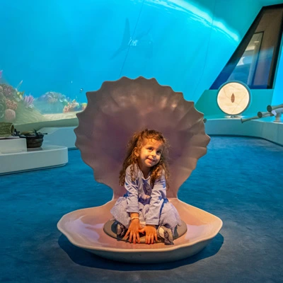 Young girl sitting playfully inside a large sculpted pearl shell in the middle of the gallery, surrounded by vivid marine-themed projections.