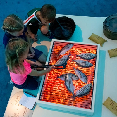 Three children roleplaying a traditional cooking activity, using tongs to place toy fish over a simulated grill lit with glowing red light to mimic fire.