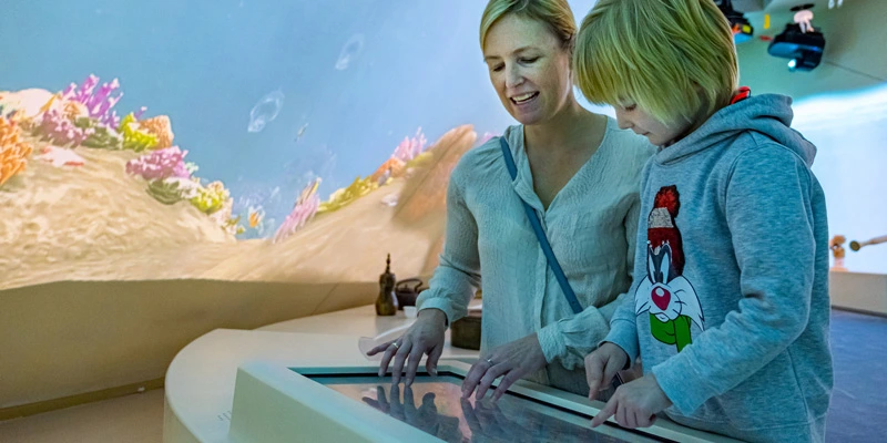 Child and adult woman exploring an interactive touch display set into a counter, with vibrant coral reef animations projected on the surrounding gallery walls.