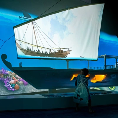 Young boy exploring a large boat model beneath a sail-shaped screen projecting a pearl diving scene, opening side compartments to discover the vessel’s interior features.