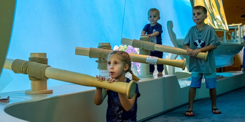Three children using large interactive wooden oars, simulating the rowing experience of pearl divers in a boat installation surrounded by an underwater environment.