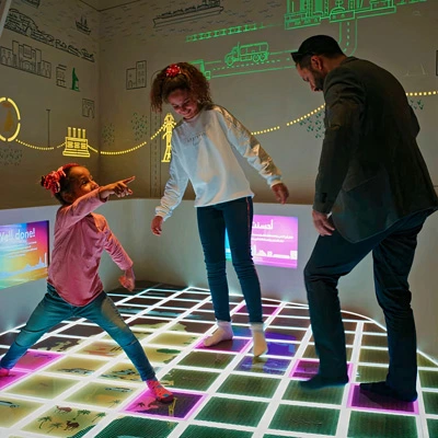 Two children and an adult joyfully interacting with an illuminated game floor, triggering projected animations and feedback related to Qatar’s energy journey.