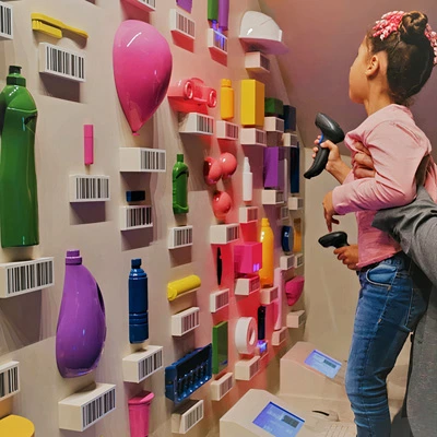 Child scanning colorful 3D-printed plastic objects shaped like consumer goods using a handheld barcode scanner at an interactive wall.
