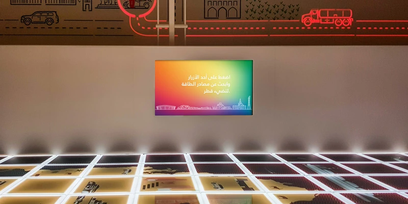 Illuminated interactive floor displaying a stylized map of Qatar, with LED-lit illustrations on the wall representing national energy sources. A rainbow-colored screen above the floor provides game instructions in Arabic.