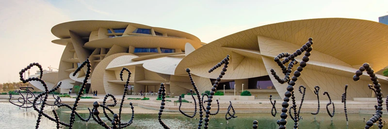 Exterior view of the National Museum of Qatar, with its iconic interlocking disc design and water features, seen through an artistic sculptural installation in the foreground.