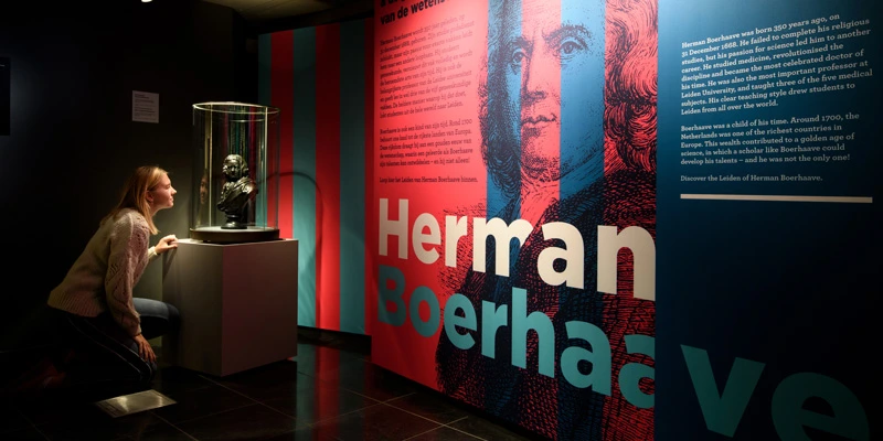 A young woman kneeling in front of a glass case containing a bronze bust, carefully observing the sculpture below a graphic wall that combines Herman Boerhaave’s name and stylized portrait.
