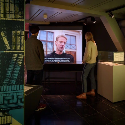Two visitors standing side by side watching a large screen playing a video interview with English subtitles, in a gallery section centered on experimentation and scientific thought.