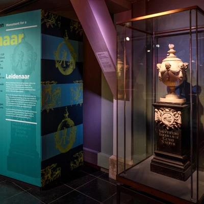 Glass case displaying a decorative urn placed on a dark pedestal, accompanied by a colorful wall panel that presents contextual information about Boerhaave’s life and work in Leiden.