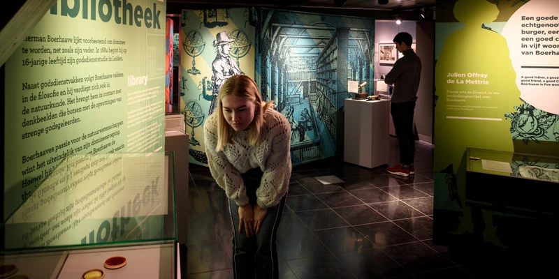 A young woman leaning forward to read a display in a room filled with colorful graphics and scientific illustrations, while a man in the background engages with another exhibit.