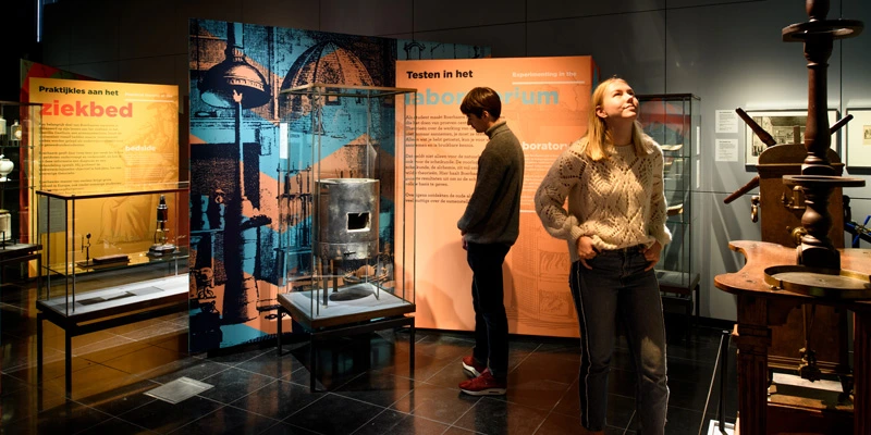 Two visitors observing a collection of scientific and medical instruments in glass cases, surrounded by vibrant graphic panels illustrating bedside practices and experimental methods.