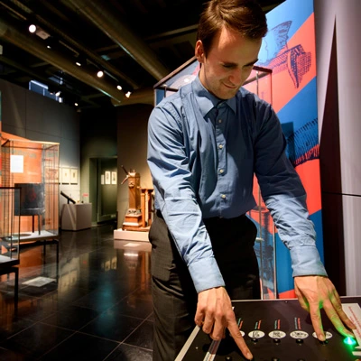 A man in a blue shirt using a tactile interactive station, with buttons and dials, in a gallery filled with historic scientific tools and instructional displays.