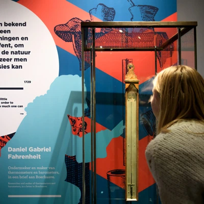 A young woman carefully examining a historical thermometer encased in glass, set against a brightly colored wall display with interpretive text about Fahrenheit’s innovations.