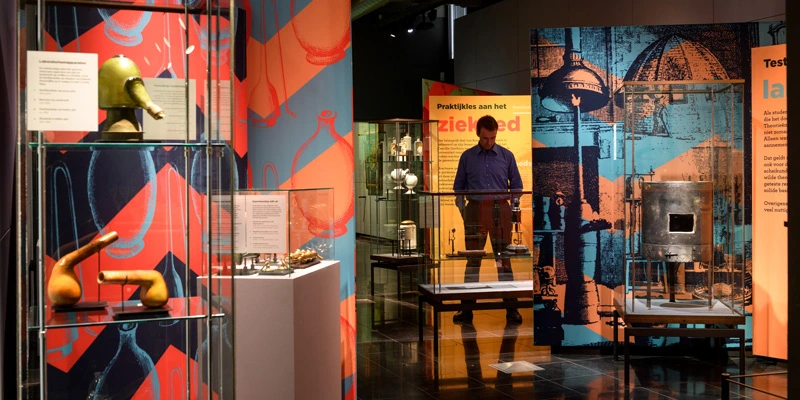 Museum visitor exploring an exhibit of glass-encased scientific instruments and apothecary tools, surrounded by stylized wall graphics referencing laboratory experimentation.