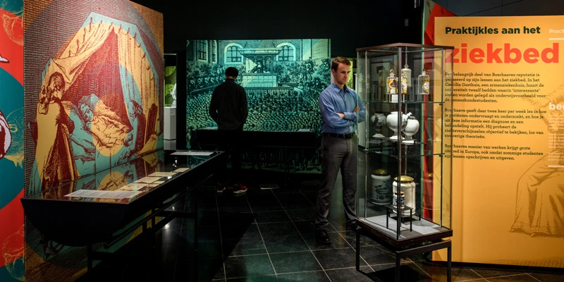 Two museum visitors navigating a gallery on bedside teaching, featuring engraved medical scenes, information panels, and historic ceramic vessels inside glass displays.