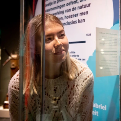 Young woman leaning over a display case to observe a historical scientific object, with detailed background illustrations and text highlighting the tool’s significance.