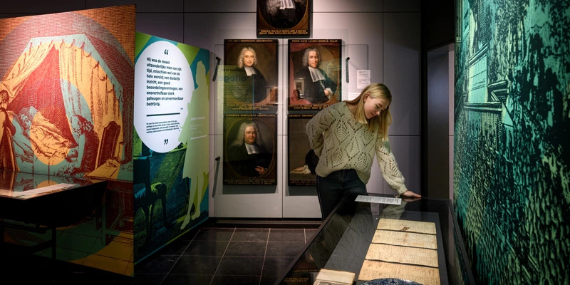Woman closely reading a glass case containing handwritten medical notes, with a backdrop of engraved portraits and a large wall illustration of a teaching scene.